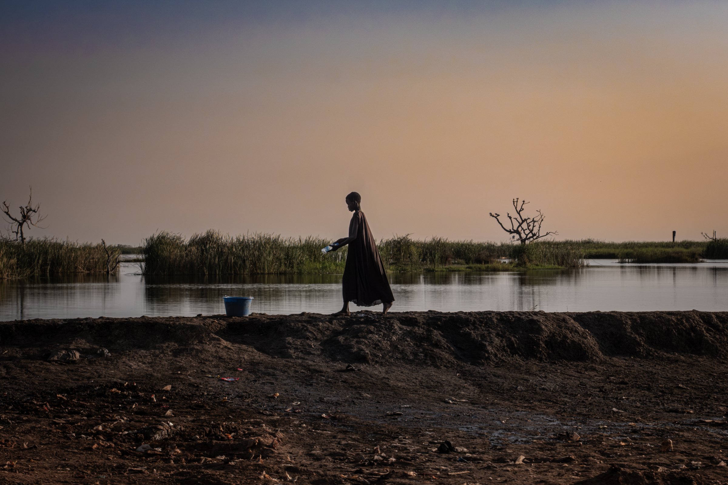 Une jeune fille marche sur l'une des digues de terre qui protègent Bentiu, une bouteille à la main. Autour d'elle, les eaux stagnantes témoignent d'un paysage transformé par des années d'inondations. État d'Unité, Soudan du Sud, mars 2026.