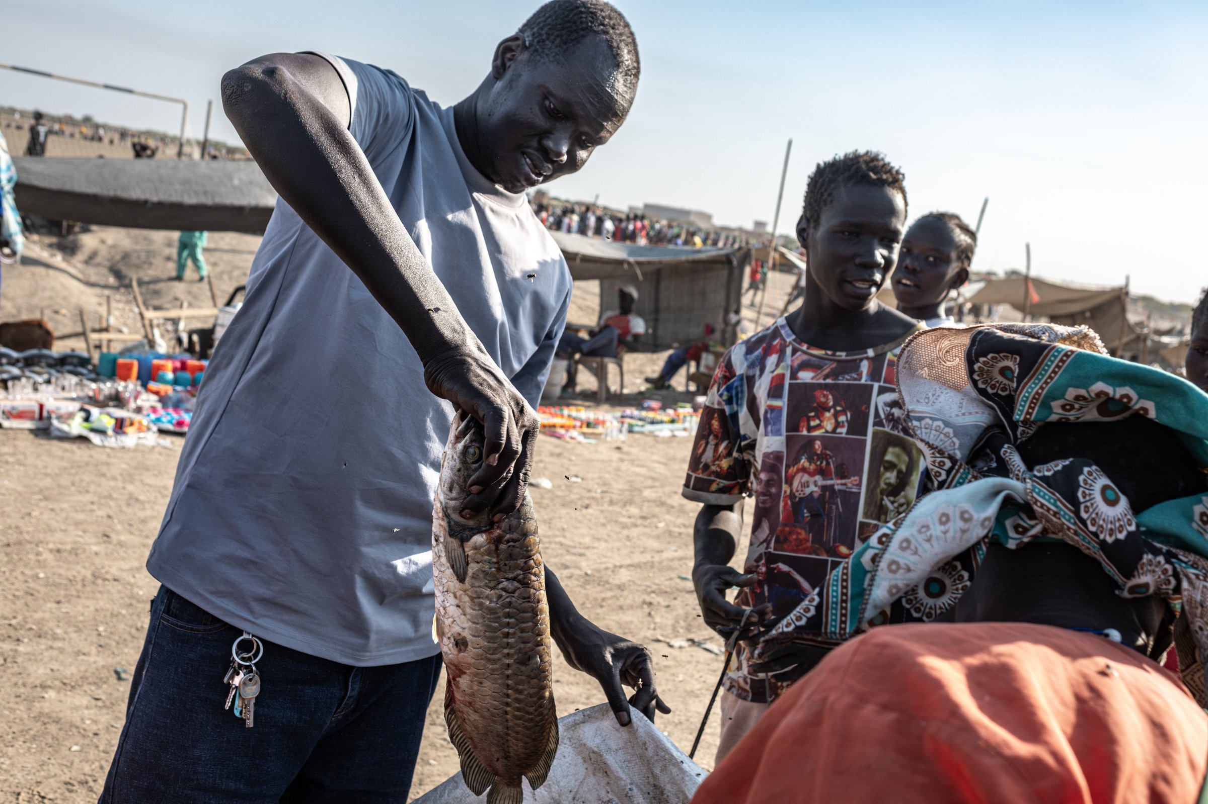 Simon Gatkuoth montre un poisson au marché du camp de déplacés de Bentiu. Comme beaucoup d'autres habitants, il affirme que certaines captures provenant des zones proches des installations pétrolières sont vendues à prix réduit parce qu'elles sont jugées contaminées. État d'Unité, Soudan du Sud, mars 2026.