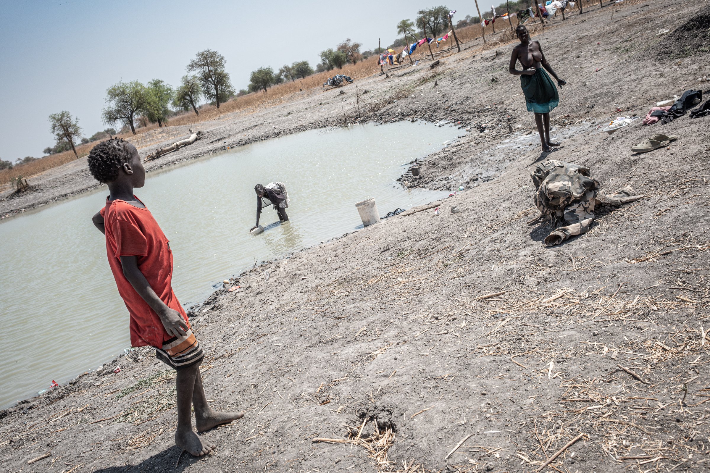 Des femmes et leurs enfants se rassemblent près d'une étendue d'eau dans une dépression de Rotriak. Selon les habitants, cet endroit faisait autrefois partie d'un bassin de rétention lié aux activités pétrolières. État d'Unité, Soudan du Sud, mars 2026.