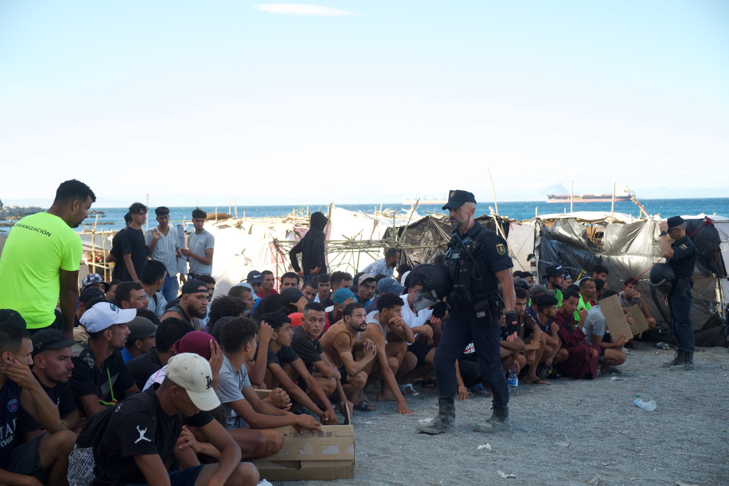 Sous la surveillance de la Police nationale, des migrants attendent, accroupis, la distribution quotidienne de nourriture.