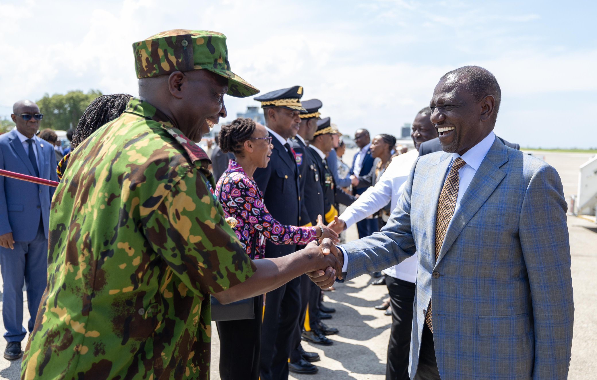 The image shows a formal scene at an airport where a group of people is gathered, likely to welcome a dignitary or a visitor. In the foreground, two men are shaking hands; one is dressed in military camouflage, and the other is in a formal suit. Around them, there are officials in uniform and business attire lined up, indicating a significant or ceremonial occasion. The setting seems to be sunny, contributing to a positive atmosphere.