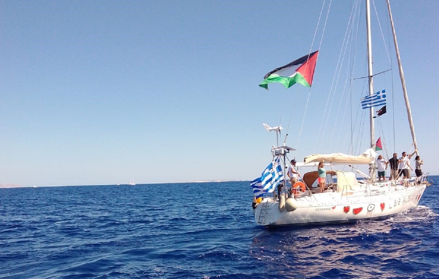 The image depicts a sailboat on calm blue waters. The boat features several flags flying from its mast: notably, a Palestinian flag, a Greek flag, and a blue-and-white striped flag typical of Greece. The sky is clear and bright, suggesting a sunny day at sea. The surrounding water appears serene, reflecting the sunlight.