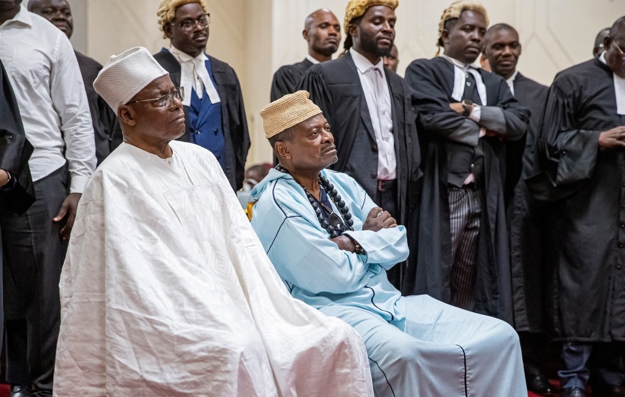 The image shows a group of individuals in formal attire, likely in a courtroom or legal setting. Two prominent figures in the foreground are seated, dressed in traditional attire. The person on the left wears a white outfit and a hat, while the individual on the right is in an aqua-colored garment with a distinctive headpiece. Behind them, several people are standing, some in legal robes, suggesting a gathering of lawyers or officials. The atmosphere appears serious and formal, indicating an important event or proceedings.