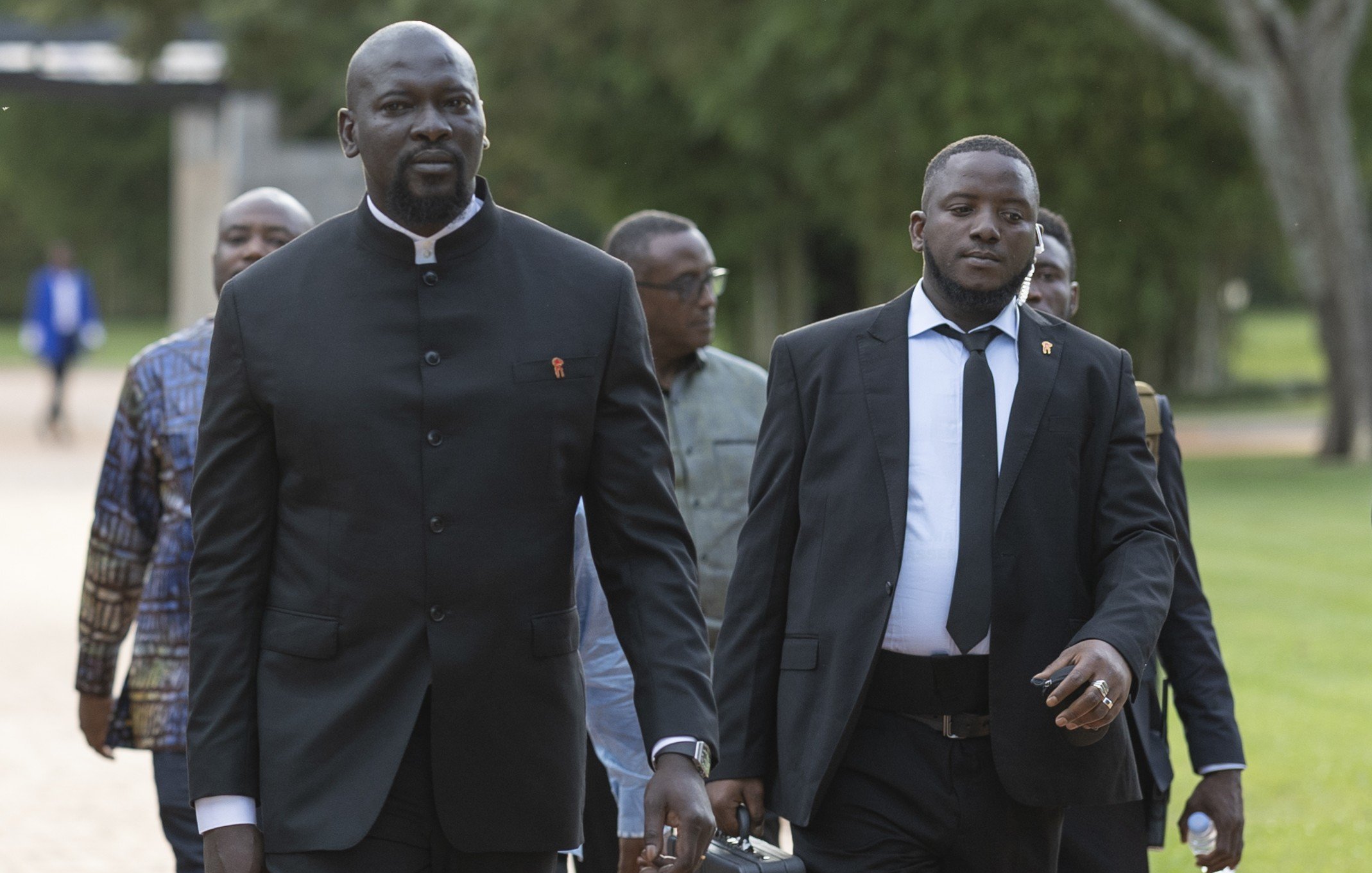 The image depicts a group of several men walking together in a park-like setting. The man in the foreground, dressed in a black suit with a high collar, appears confident and focused. He is accompanied by another man wearing a black suit and tie, who carries a briefcase. The background features lush greenery, suggesting an outdoor event or gathering. Other men can be seen following closely behind, contributing to a formal yet relaxed atmosphere.