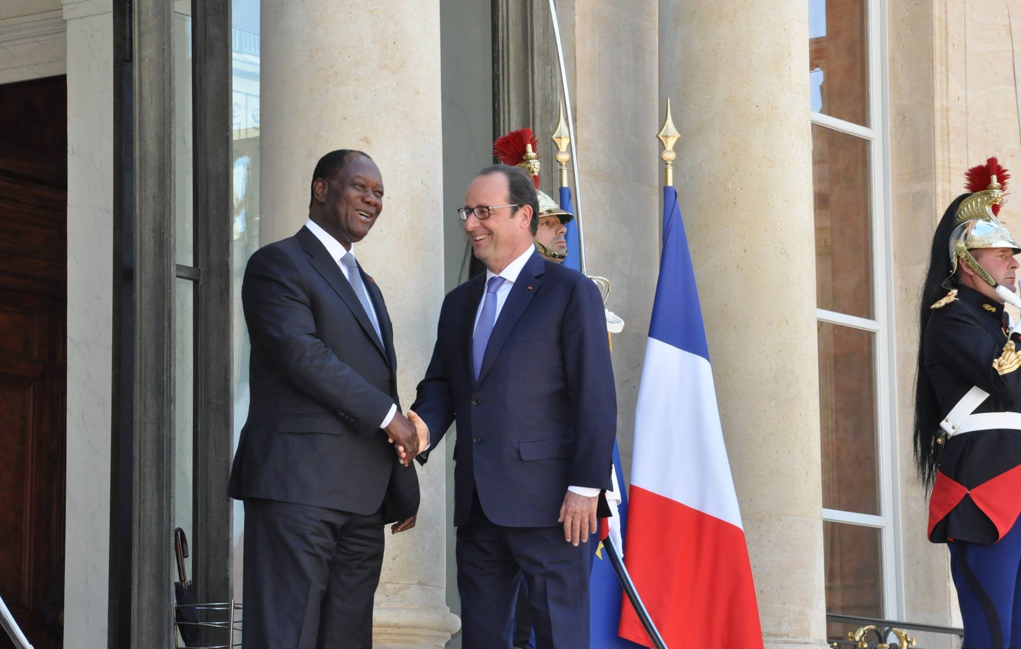 The image shows two men shaking hands outside a large building, likely a government or official residence. One man is wearing a dark suit and the other is dressed in a suit and tie, likely representing different nations. Behind them, there is a large flag of France, and guards in traditional uniforms stand watch. The setting suggests a formal meeting or diplomatic engagement.