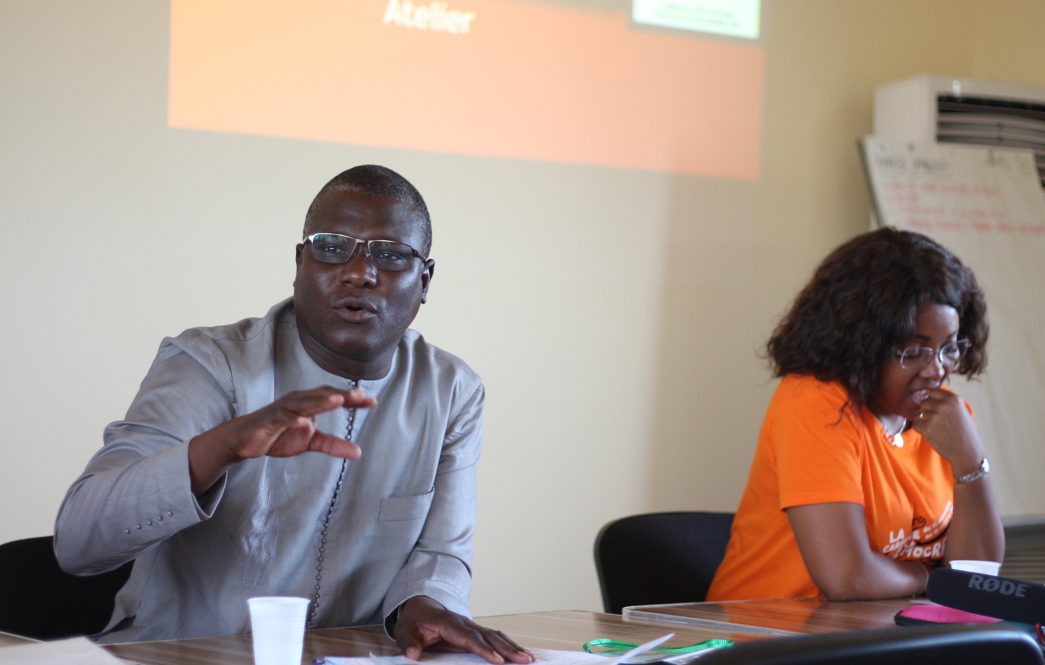 L'image représente un atelier se déroulant dans une salle de réunion. Au premier plan, deux personnes sont assises à une table. À gauche, un homme portant une chemise traditionnelle grise. Il semble s'exprimer avec enthousiasme, levant une main pour souligner un point important. À droite, une femme avec des cheveux bouclés est assise, portant un t-shirt orange. Elle l'écoute attentivement. En arrière-plan, un écran présente le mot "Atelier" en lettres d'origine orange. La scène est empreinte d'une atmosphère collaborative et interactive.