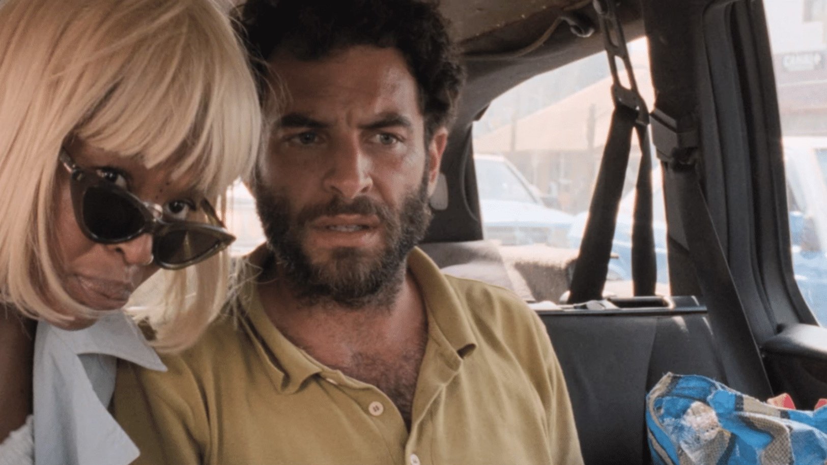 The image shows two people inside a vehicle. One person, likely wearing a blonde wig and sunglasses, leans towards the other, who has a beard and a concerned expression. The setting appears casual and slightly cramped, with a background that suggests they are parked in a busy area. The overall mood seems tense or serious, as both individuals have focused looks on their faces.