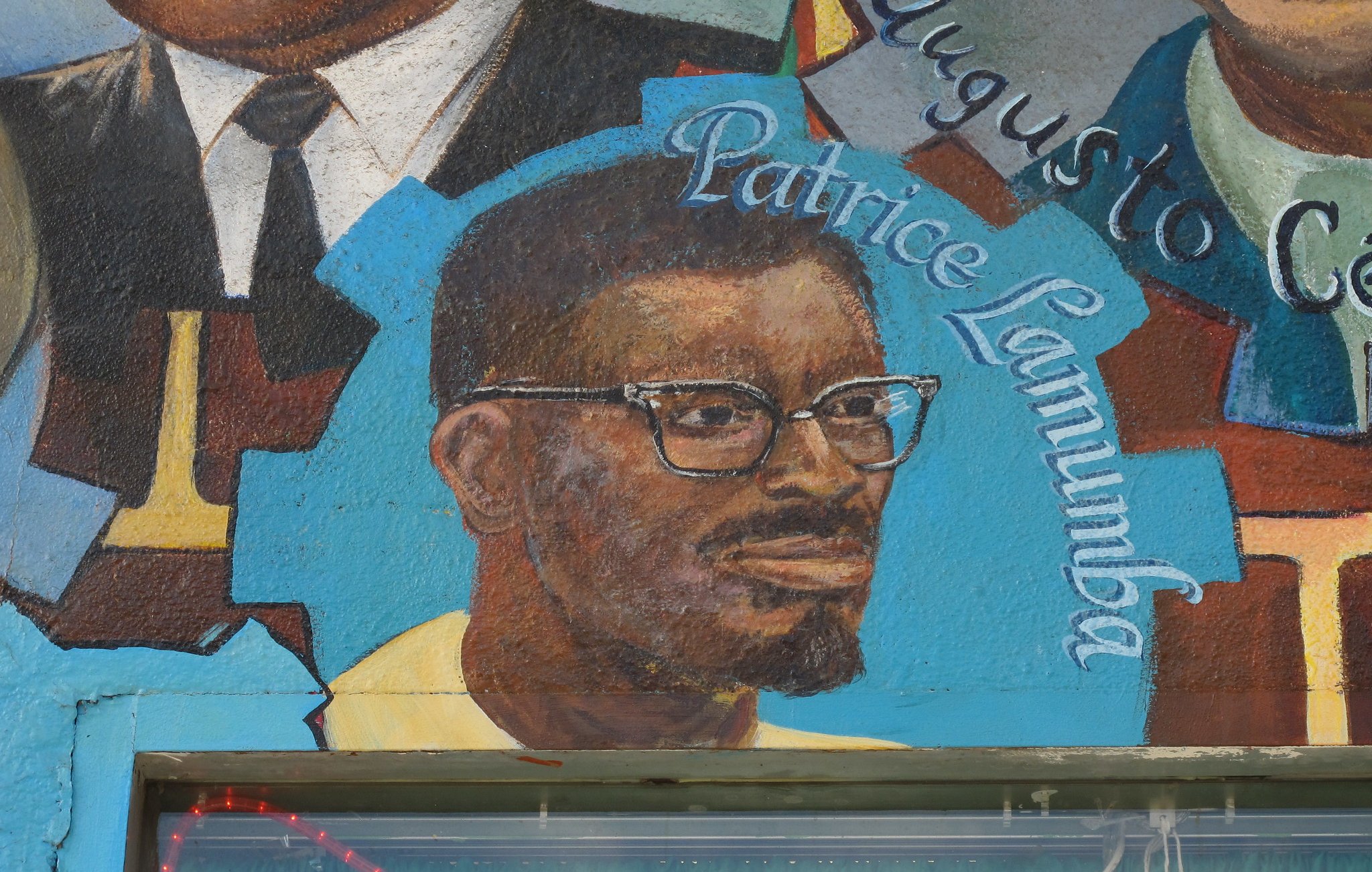 The image features a mural that includes a portrait of a man with glasses and a beard, identified as Patrice Lumumba. He is depicted in a colorful artistic style, wearing a yellow shirt. The mural may also include other figures or elements, but this particular part focuses on Lumumba's likeness alongside his name, highlighting his significance. The background is vibrant, contributing to the overall visual impact of the mural.