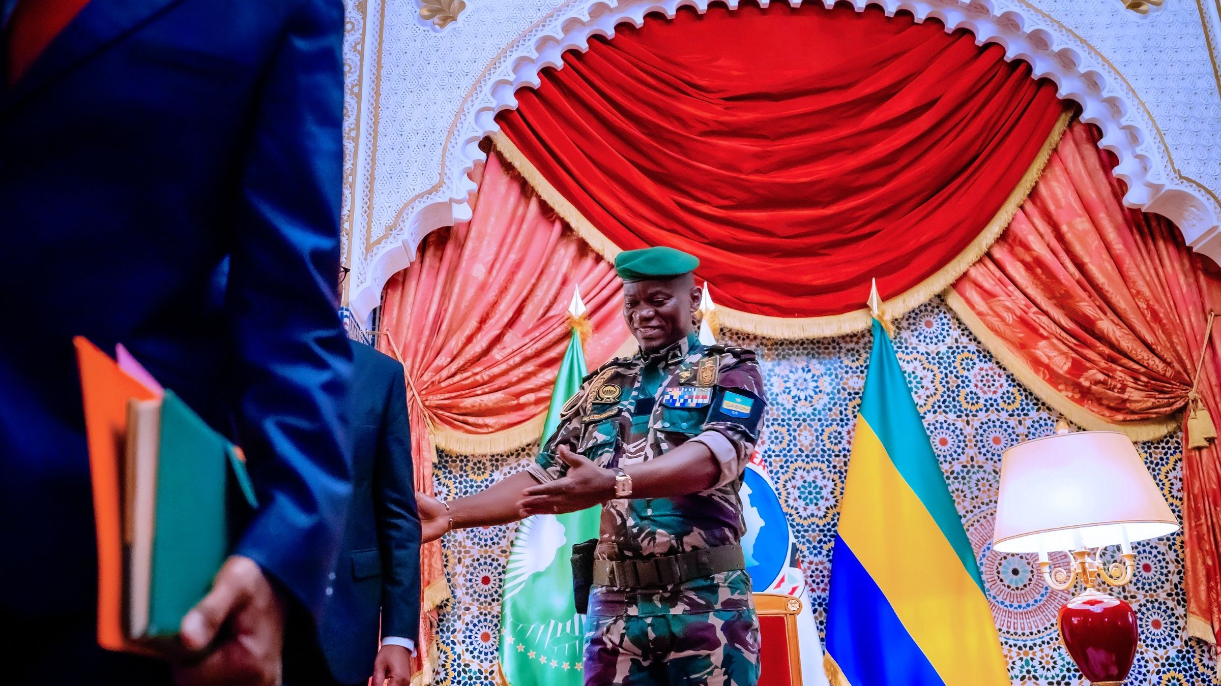The image shows a scene set in an ornate room. A military officer, dressed in camouflage with a green beret, is smiling and appears to be interacting with another individual. The background features decorative elements, including curtains and flags, likely representing Gabon or an official entity. There are also books and documents in the hands of a person standing to the left, indicating a formal context, possibly a meeting or ceremony. The overall atmosphere seems official and ceremonial.