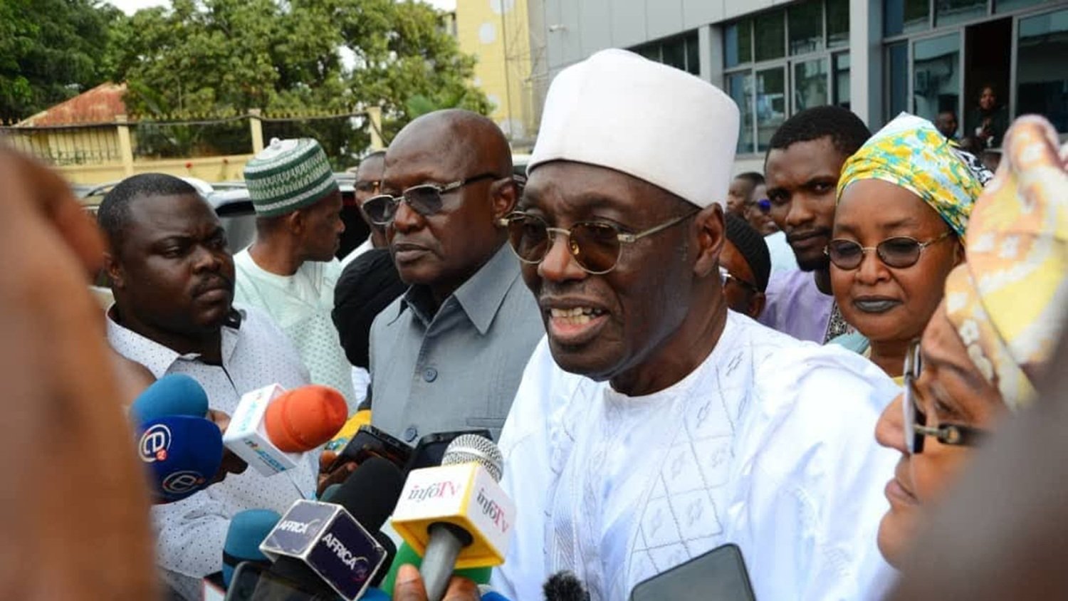 The image shows a group of people gathered around a man wearing a white traditional outfit and a white cap. He appears to be speaking to the media, as indicated by the microphones and cameras directed at him. Surrounding him are several other individuals, some of whom are dressed in formal attire, while others wear traditional garments. The background features a building, and the atmosphere suggests a press event or a public announcement.