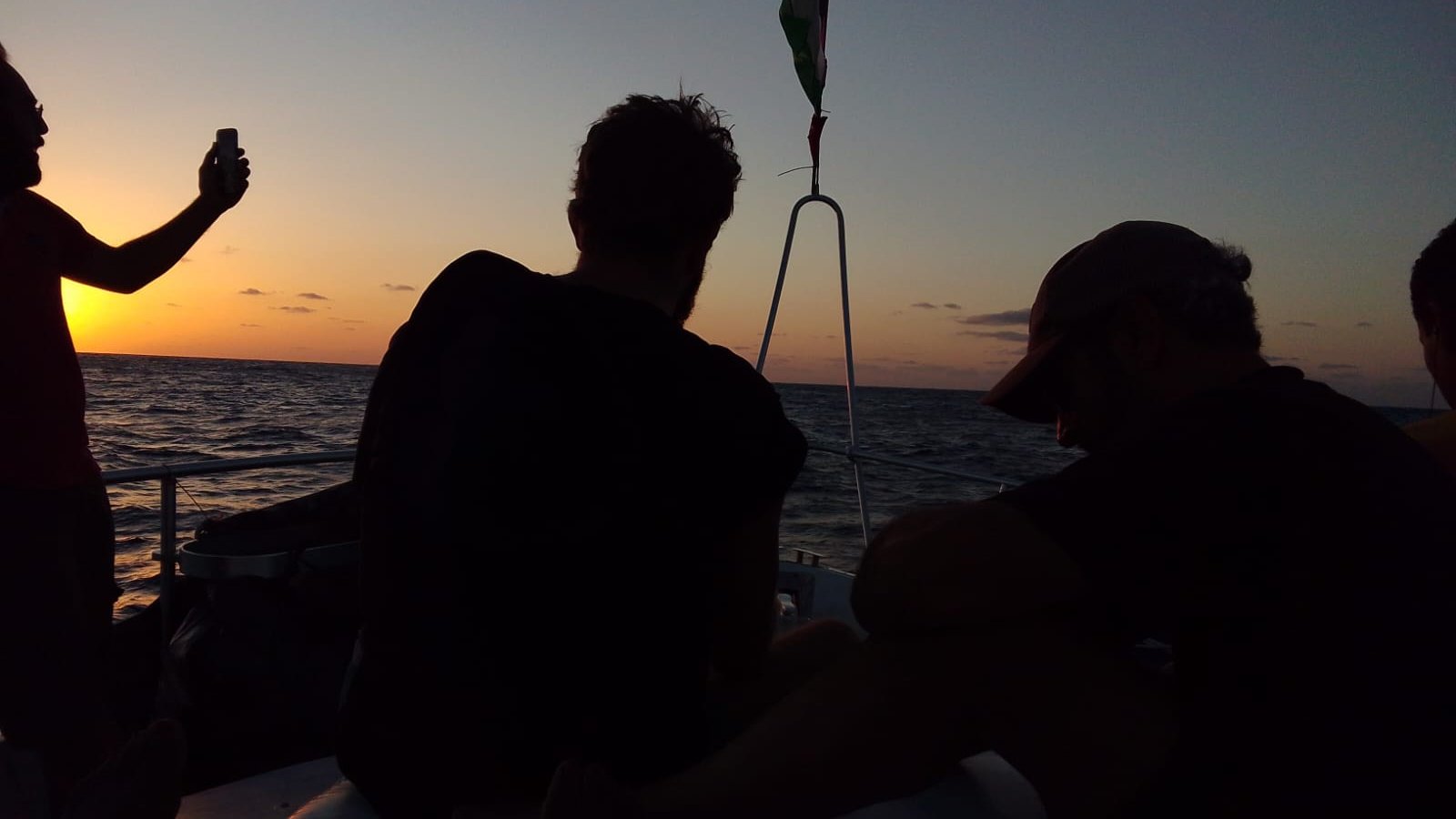 The image shows a group of people on a boat at sunset. The sky is painted in warm hues of orange and yellow as the sun sets on the horizon. Two people are visible, sitting with their backs to the camera, seemingly enjoying the view. One person is standing and holding a phone, possibly taking a photo. The water appears calm, reflecting the colors of the sky. The overall atmosphere is serene and relaxed.