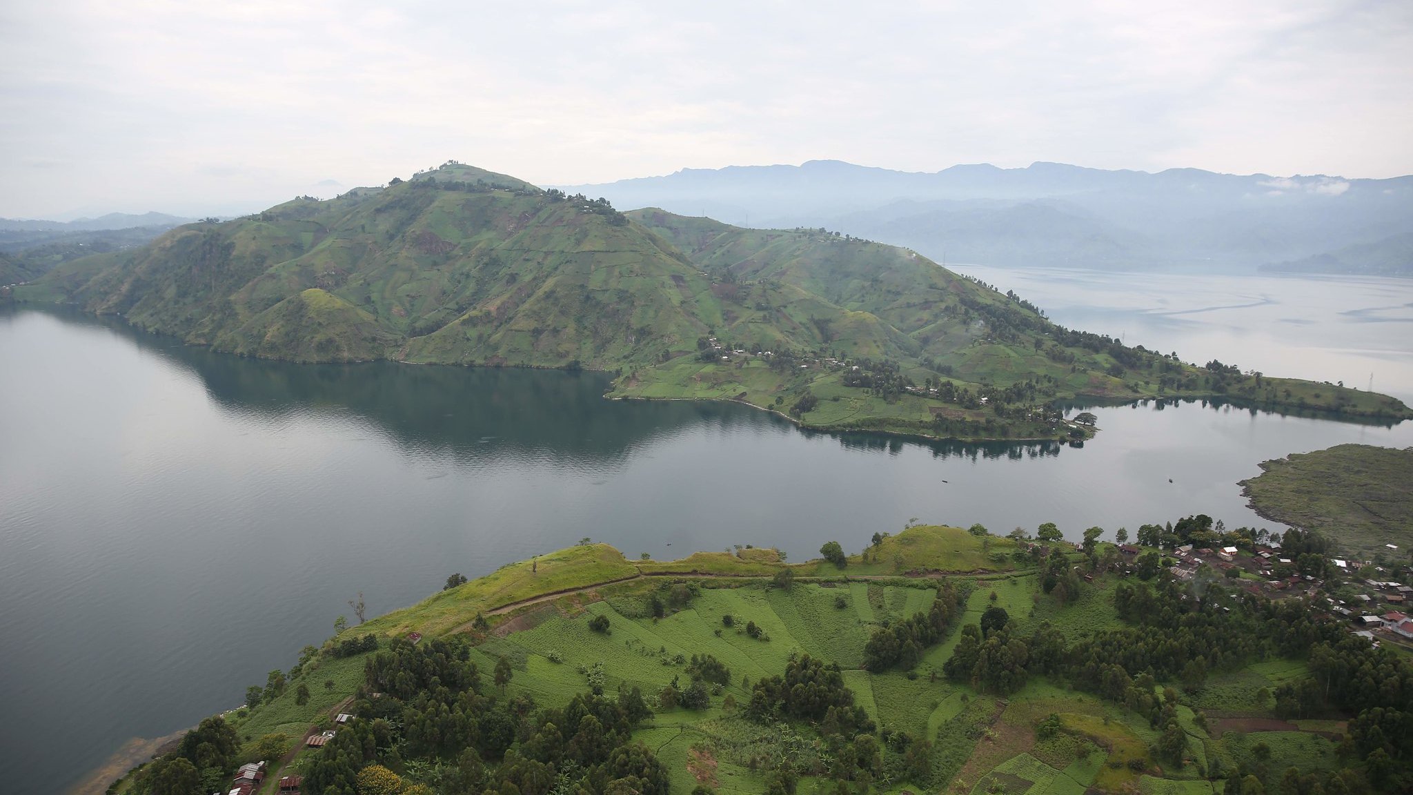 The image showcases a breathtaking landscape featuring lush green hills and a large lake. The hills are gently sloped and dotted with patches of vegetation, indicating a fertile area. In the foreground, there are scattered houses or structures, while the calm waters of the lake reflect the surrounding scenery. In the background, more mountainous terrain rises, adding depth to the view under a slightly overcast sky. The overall atmosphere is serene and picturesque, highlighting the natural beauty of the region.