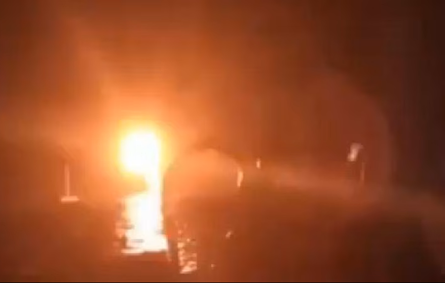 The image appears to show a bright light source in a dark environment, suggesting a fire or an explosion. The intense glow creates a dramatic contrast against the dark background, and there are vague silhouettes or shapes surrounding the light, which could indicate the presence of objects or people nearby. The overall atmosphere seems tense and chaotic due to the brightness and obscurity of the other elements in the scene.