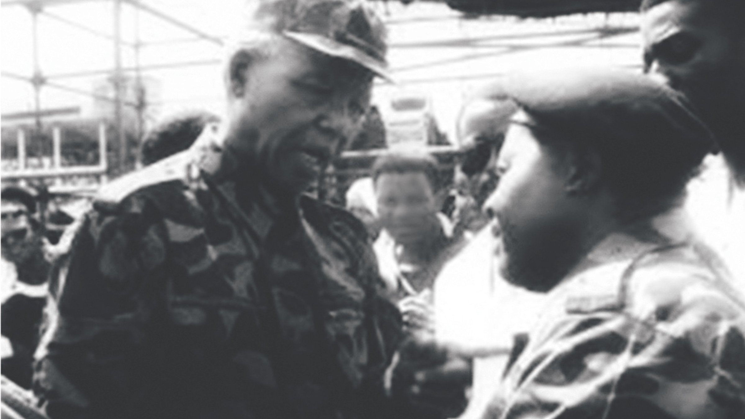 The image appears to be a historic black-and-white photograph depicting two men engaged in a conversation. One man is wearing military camouflage and appears to be in a position of authority, while the other is also dressed in military attire. They are surrounded by a group of people, indicating a significant setting, possibly a public event or gathering. The expressions on their faces suggest a serious discussion. The context hints at a moment of political or military importance, likely related to South Africa's history.