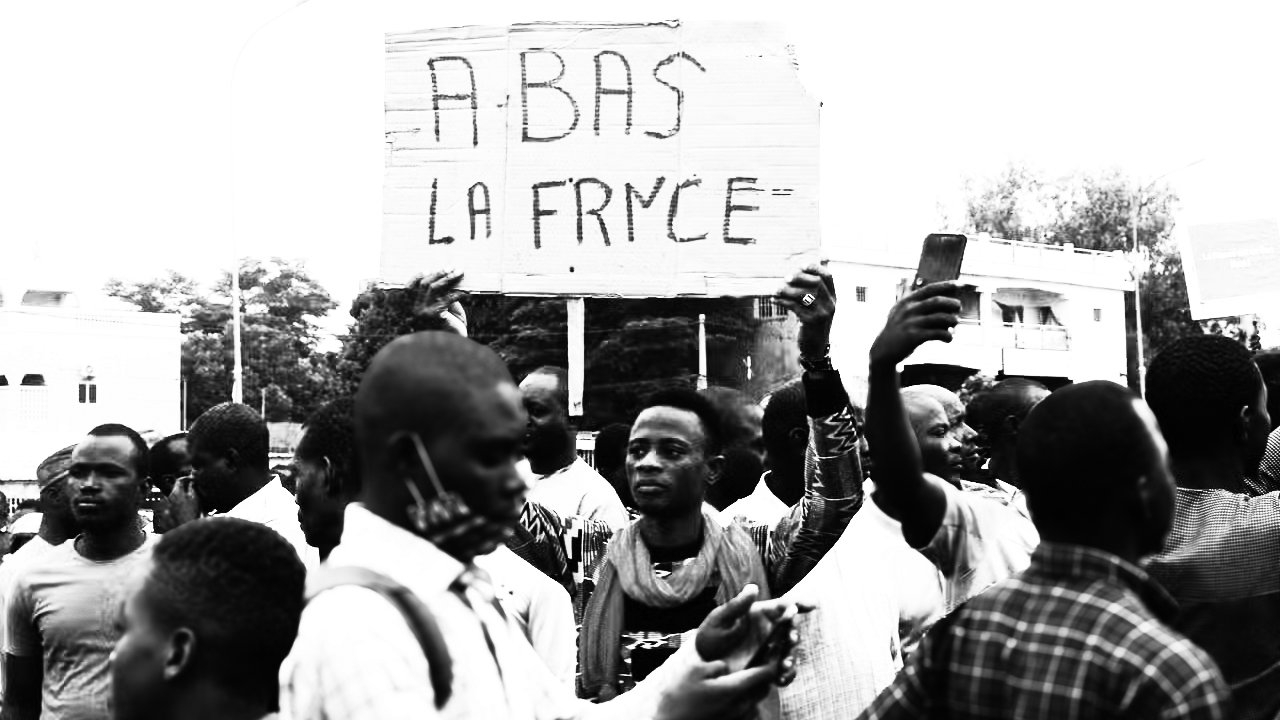 L'image représente une scène de manifestation. Au premier plan, plusieurs personnes se rassemblent, certaines levant les bras. Un homme, au centre, tient une grande pancarte avec l'inscription "ABAS LA FRYCE" en lettres capitales. La foule semble engagée et énergique, exprimant une certaine frustration ou un message de contestation. L'ambiance est chargée d'émotion, avec des visages déterminés et des gestes expressifs, dans un cadre urbain qui suggère un lieu public. Les vêtements des manifestants varient, ajoutant à la diversité de la scène.