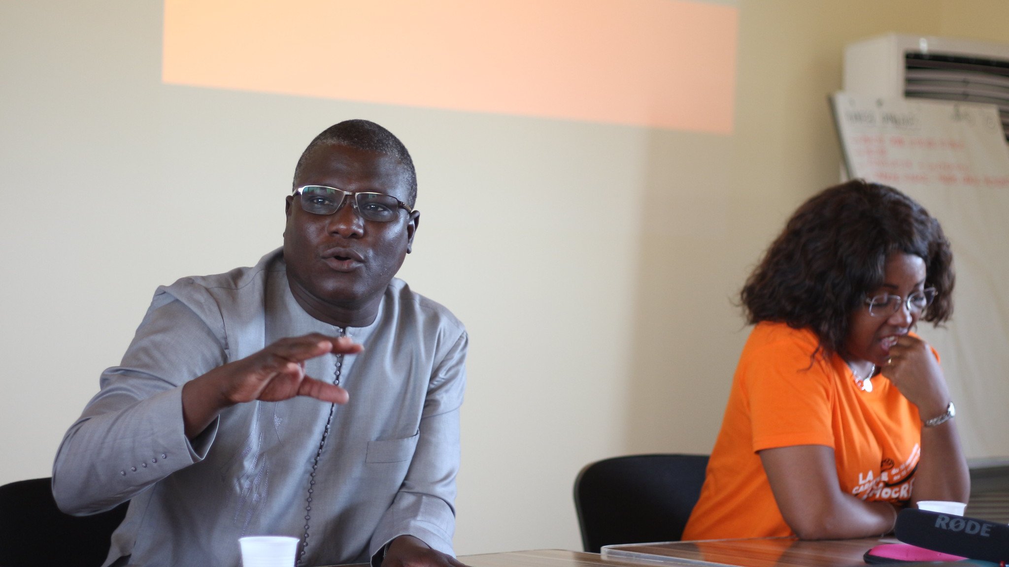 L'image représente un atelier se déroulant dans une salle de réunion. Au premier plan, deux personnes sont assises à une table. À gauche, un homme portant une chemise traditionnelle grise. Il semble s'exprimer avec enthousiasme, levant une main pour souligner un point important. À droite, une femme avec des cheveux bouclés est assise, portant un t-shirt orange. Elle l'écoute attentivement. En arrière-plan, un écran présente le mot "Atelier" en lettres d'origine orange. La scène est empreinte d'une atmosphère collaborative et interactive.