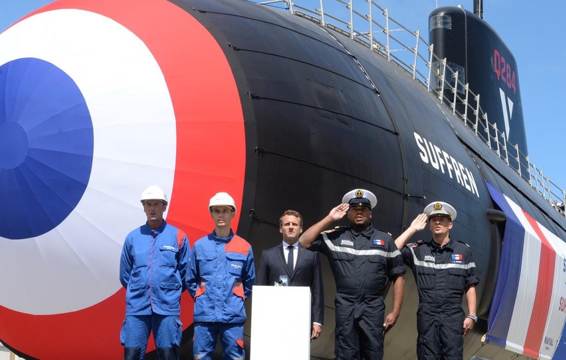 L'image représente un sous-marin de type Suffren, mis en avant lors d'une cérémonie en extérieur. Le sous-marin, aux couleurs caractéristiques bleu, blanc et rouge, se dresse majestueusement avec une grande coque stylisée. Le chiffre « 12 » est visible sur le côté. Devant le sous-marin, on voit plusieurs personnes en tenue de travail. À gauche, deux hommes portent des combinaisons bleues, un troisième homme, en costume, semble féliciter le projet, tandis que le quatrième, en uniforme, adopte une posture militaire. L'arrière-plan est dégagé avec un ciel bleu, ce qui donne une impression de journée ensoleillée. La scène évoque un moment de fierté et de célébration pour les employés et les responsables militaires, symbolisant un jalon important dans le domaine naval.