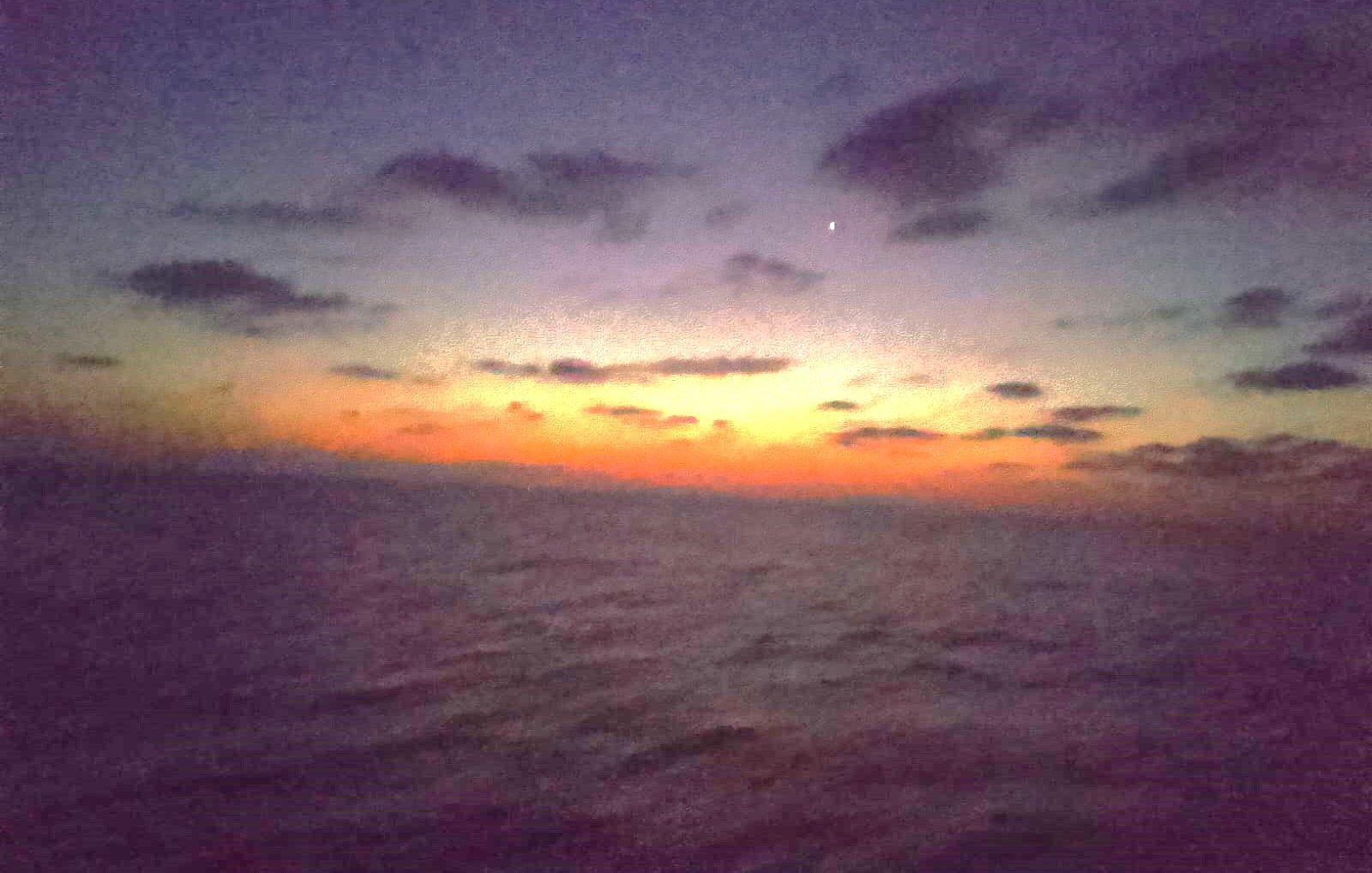 The image shows a serene seascape during twilight. The sky is transitioning from daylight to night, displaying a gradient of colors ranging from deep blue to orange near the horizon. Clouds are scattered across the sky, adding to the atmospheric depth. A single bright star is visible in the upper right portion of the image, contributing to the tranquil mood of the scene. The calm ocean reflects these colors, creating a peaceful and beautiful setting.
