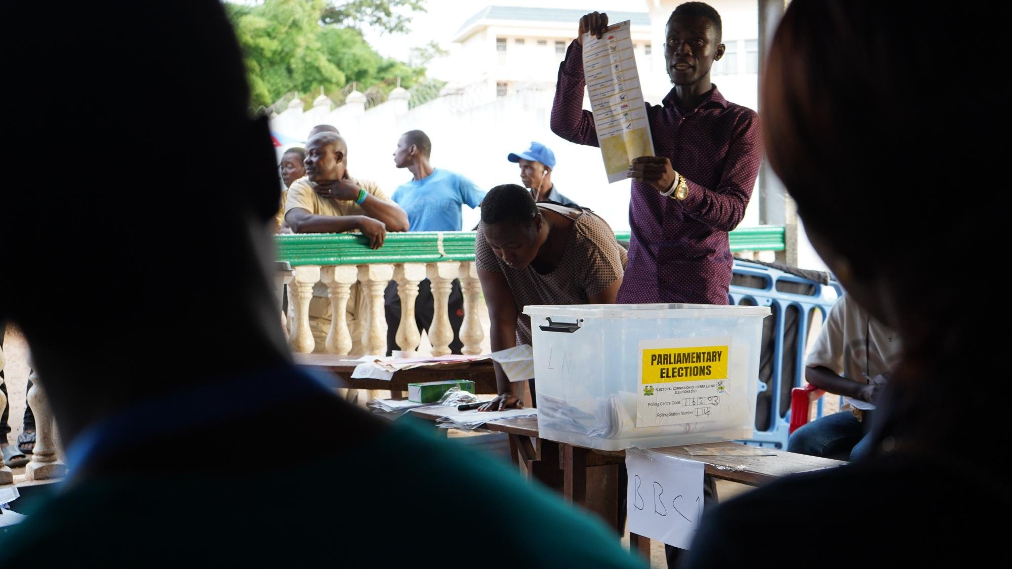 The image depicts a scene from a polling station during a parliamentary election. A man, likely an election official, is holding up a ballot sheet for voters to see. In front of him is a transparent box labeled "PARLIAMENTARY ELECTIONS," suggesting it's used for collecting votes. In the background, several people are gathered, observing the process, while some focus on the official. The setting appears to be outdoors, possibly in a community area, and there's a sense of engagement and participation in the democratic process.