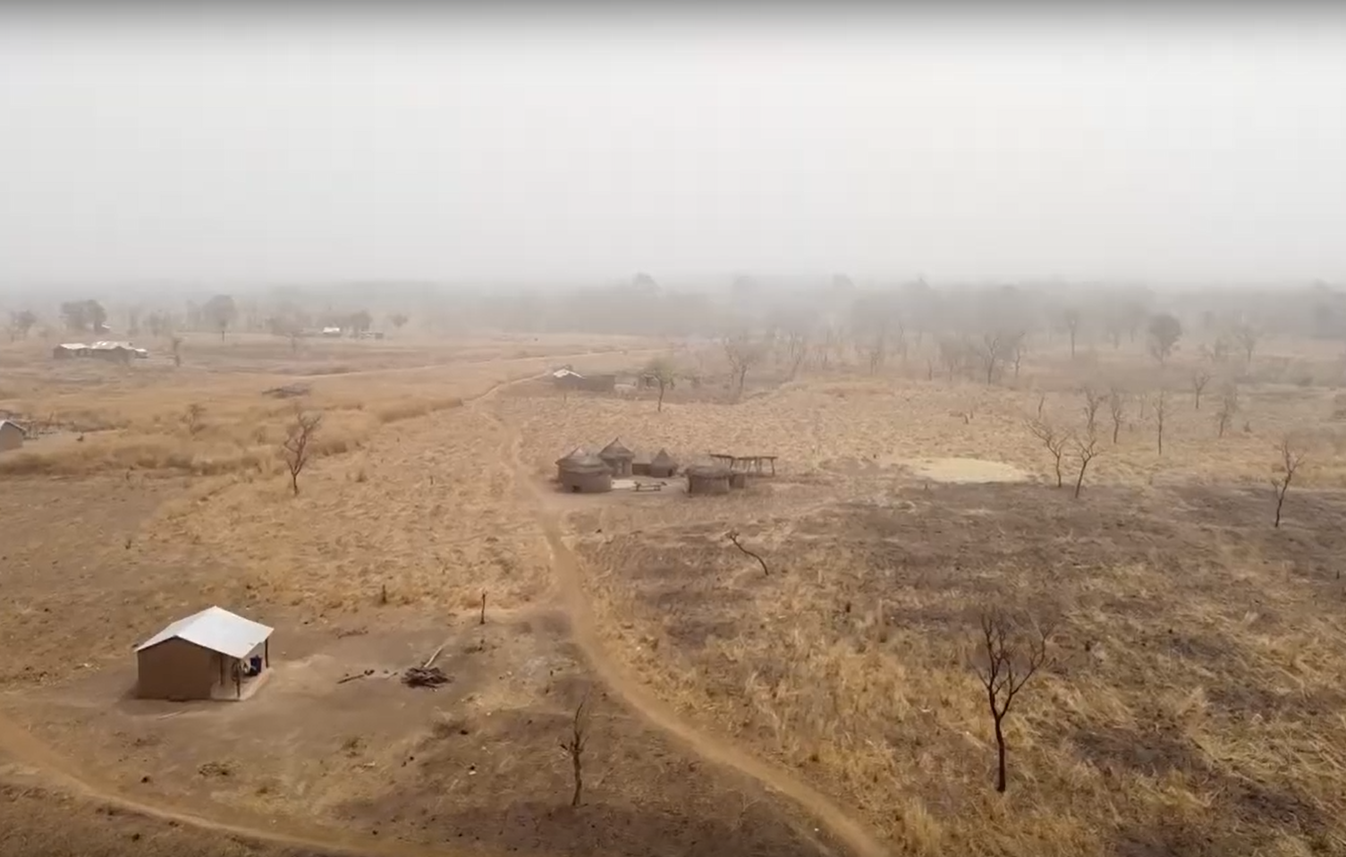 The image depicts a rural landscape characterized by dry, barren land. There are patches of brown grass and sparse vegetation, indicating an arid environment. In the foreground, a small structure with a white roof sits alongside a dirt path. In the background, a few more buildings can be seen, scattered amidst the landscape. The atmosphere appears hazy, possibly due to dust or smoke, giving the scene a muted and subdued appearance. Sparse trees dot the area, and the overall setting suggests a remote, pastoral region.