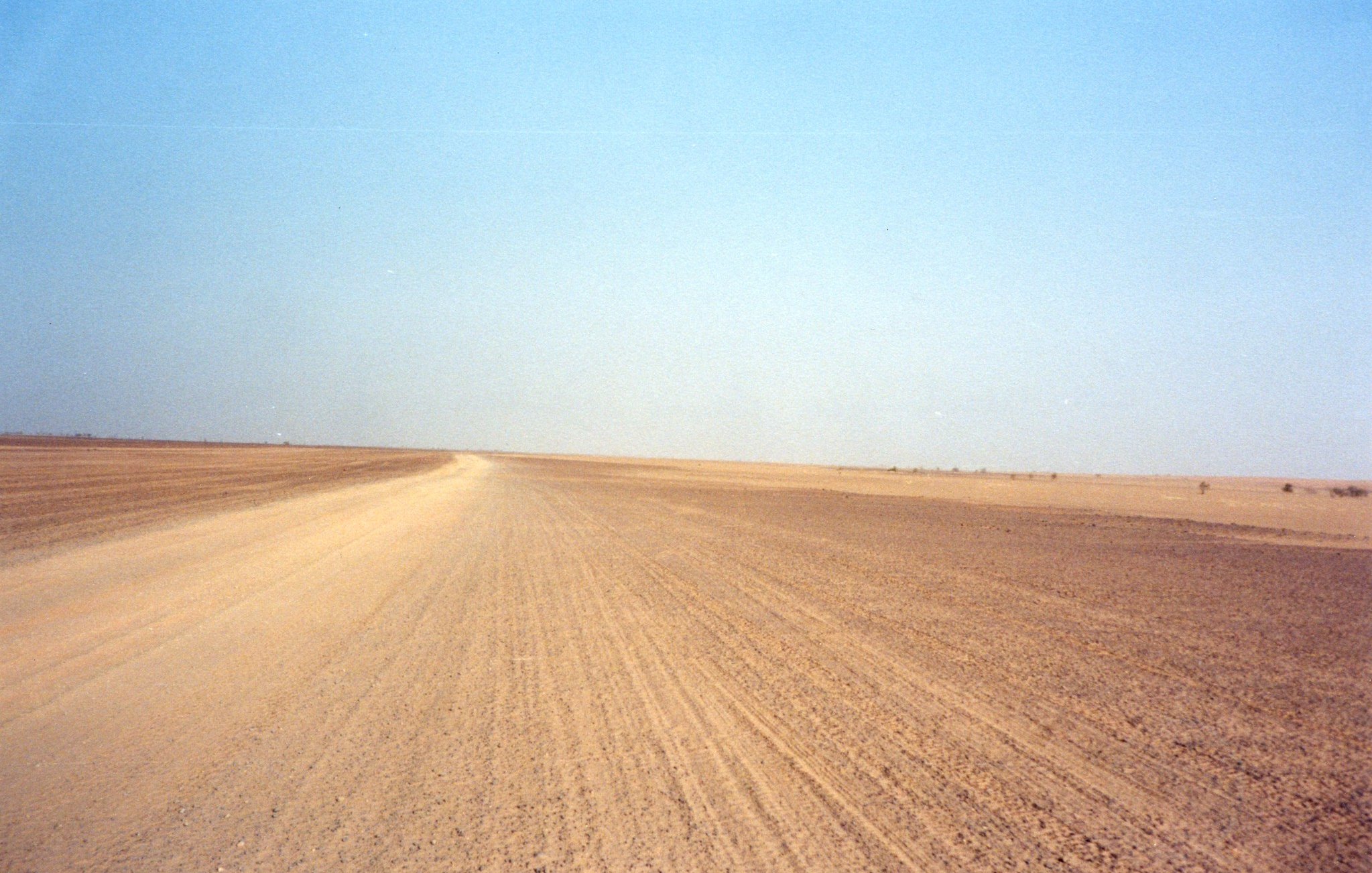 The image depicts a barren landscape characterized by a vast, empty stretch of land, likely a desert or arid region. The foreground features a dirt road that leads into the distance, flanked by dry, flat terrain. The sky overhead is clear and blue, creating a stark contrast with the earthy tones of the ground. The overall scene conveys a sense of isolation and openness, typical of remote, dry environments.