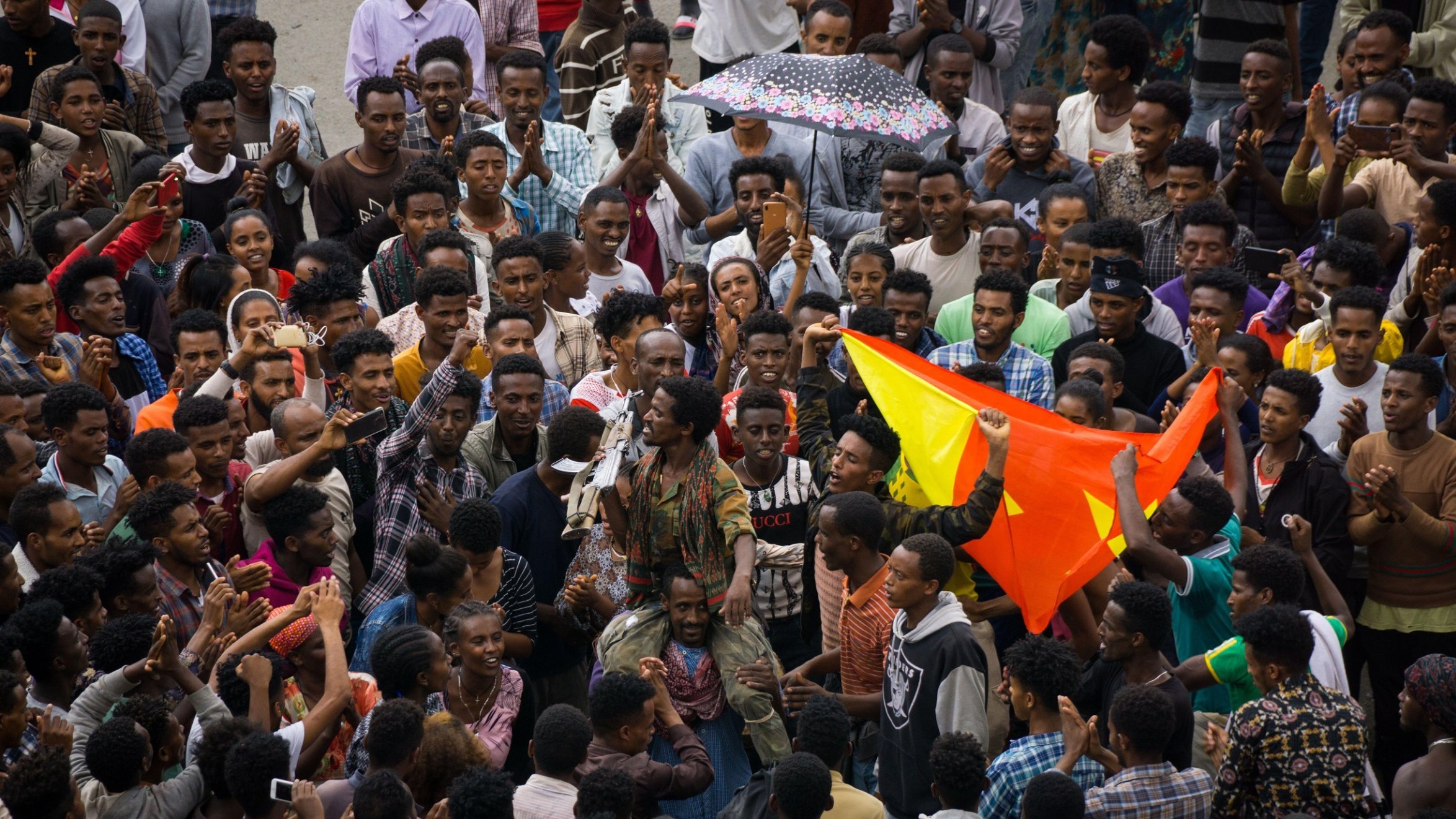 The image depicts a large gathering of people in a vibrant setting, likely representing a celebration or demonstration. Crowds are tightly packed together, with some individuals holding colorful flags, possibly indicating a cultural or political event. One person stands out in the center, engaging the crowd, which looks enthusiastic and expressive. The mood appears lively, with a sense of unity among the participants. Various expressions and clothing styles suggest a diverse group, reflecting a shared purpose or message. An umbrella can be seen, hinting at the weather conditions. Overall, it captures a moment of collective energy and communal spirit.