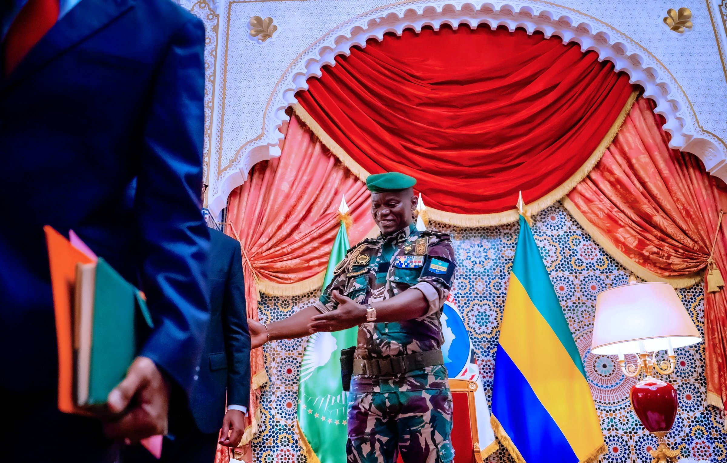 The image shows a scene set in an ornate room. A military officer, dressed in camouflage with a green beret, is smiling and appears to be interacting with another individual. The background features decorative elements, including curtains and flags, likely representing Gabon or an official entity. There are also books and documents in the hands of a person standing to the left, indicating a formal context, possibly a meeting or ceremony. The overall atmosphere seems official and ceremonial.
