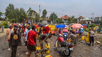 The image depicts a crowded scene in an outdoor setting, likely a market or gathering place. People are seen collecting water from yellow jerry cans, some of which are stacked nearby. The atmosphere appears busy and somewhat chaotic, with individuals interacting and engaging in various activities. There are motorcycles and umbrellas visible, suggesting a warm, sunny environment. The ground is littered with debris, indicating a lack of cleanliness in the area. The background features buildings and trees, contributing to the urban landscape.