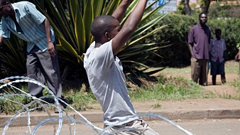 The image depicts a scene where a young man is on his knees, appearing to be in a moment of distress or surrender. He is surrounded by coils of barbed wire on the ground. In the background, there are several individuals observing the situation, some appearing to be standing casually while others are more focused on what is happening. The setting looks to be an outdoor area, possibly a street, with greenery in the background. The overall atmosphere seems tense and conveys a sense of conflict or protest.