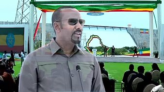 The image appears to show a man speaking at a ceremonial or public event outdoors, likely in Ethiopia. He is wearing sunglasses and has a serious expression. The background features a large stage, decorated with colorful flags, possibly representing Ethiopia. There is a crowd of people seated in front of the stage, indicating that this is a significant gathering. The event seems to be celebratory and possibly related to national pride or development.