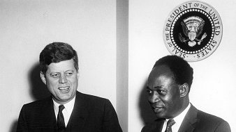 The image features two men, one of whom is John F. Kennedy, the President of the United States, and the other is likely Kwame Nkrumah, the first President of Ghana. They appear to be at a press conference or a formal meeting, with microphones in front of them. Behind them, there is a seal that represents the office of the President of the United States. Both men are dressed formally, with Kennedy in a suit and tie, and they are smiling, suggesting a cordial atmosphere.