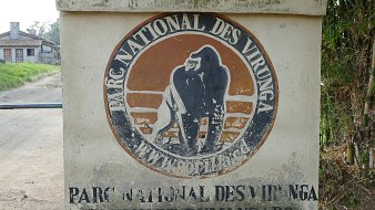 The image features a sign marking the entrance to the Virunga National Park. The sign displays the park's name in both French and English, showcasing the emblem of the park, which includes an illustration of a gorilla. The surroundings seem to depict a rural area, with a road leading away from the entrance and a building visible in the background. The overall atmosphere suggests an area of natural beauty and wildlife conservation.