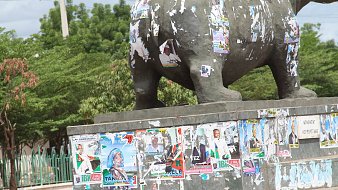 The image depicts a statue, likely an elephant, that is visibly weathered and covered in numerous political posters and flyers. These posters, in various colors, are peeling and overlapping, suggesting a history of political campaigns or events. The statue is situated in an outdoor area, surrounded by greenery and a fence in the background. The overall scene conveys a sense of urban life and the passage of time through the accumulation of posters.