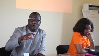 L'image représente un atelier se déroulant dans une salle de réunion. Au premier plan, deux personnes sont assises à une table. À gauche, un homme portant une chemise traditionnelle grise. Il semble s'exprimer avec enthousiasme, levant une main pour souligner un point important. À droite, une femme avec des cheveux bouclés est assise, portant un t-shirt orange. Elle l'écoute attentivement. En arrière-plan, un écran présente le mot "Atelier" en lettres d'origine orange. La scène est empreinte d'une atmosphère collaborative et interactive.