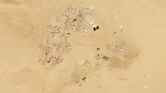 The image appears to be an aerial view of a desert landscape featuring two distinct areas. On the left side, there are ruins or archaeological sites, indicated by clusters of structures, possibly remnants of ancient buildings. Nearby, there is an airport and some roads connecting the areas. On the right side, there is a more densely populated area, showing what looks like a town or settlement named "Antic," characterized by a grid-like pattern, suggesting regular streets and organized layout. Surrounding the settlement, there are patches of greenery, likely indicating agricultural activity. The vast surrounding landscape is primarily sandy and barren, typical of desert regions.
