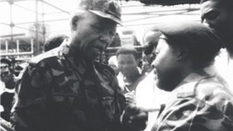 The image appears to be a historic black-and-white photograph depicting two men engaged in a conversation. One man is wearing military camouflage and appears to be in a position of authority, while the other is also dressed in military attire. They are surrounded by a group of people, indicating a significant setting, possibly a public event or gathering. The expressions on their faces suggest a serious discussion. The context hints at a moment of political or military importance, likely related to South Africa's history.