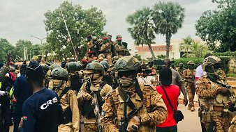 The image depicts a scene of armed soldiers in a public area. The soldiers are wearing camouflage uniforms and helmets, some with face coverings, and are equipped with rifles. In the background, there are additional soldiers on a vehicle. The atmosphere appears to be tense, with a gathering of civilians present as well, some taking photographs. There are trees and buildings in the background, suggesting an urban setting. Overall, the scene conveys a strong military presence.