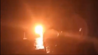 The image appears to show a bright light source in a dark environment, suggesting a fire or an explosion. The intense glow creates a dramatic contrast against the dark background, and there are vague silhouettes or shapes surrounding the light, which could indicate the presence of objects or people nearby. The overall atmosphere seems tense and chaotic due to the brightness and obscurity of the other elements in the scene.