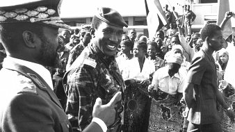 The image depicts a historical scene likely from a public event or rally. In the foreground, two men are engaging in conversation; one is dressed in a military uniform, while the other wears a military-style jacket with insignia. They both seem to be smiling and interacting positively. Behind them, a crowd of people of various ages is visible, with many holding flags and displaying enthusiastic gestures. The atmosphere appears lively, suggesting a moment of celebration or political significance. The image is in black and white, adding to its historical context.