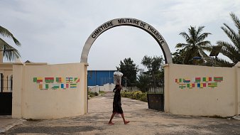 The image shows a person walking through the entrance of "Cimetière Militaire de Thiaroye," a military cemetery. The archway is prominently displayed, with the name of the cemetery written above. On either side of the entrance, there are colorful flags painted on the walls. The person in the foreground is carrying a large container on their head, and the scene is partly illuminated by natural light, surrounded by palm trees and greenery. The sky is overcast, giving a muted ambiance to the setting.