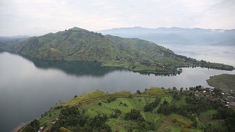 The image showcases a breathtaking landscape featuring lush green hills and a large lake. The hills are gently sloped and dotted with patches of vegetation, indicating a fertile area. In the foreground, there are scattered houses or structures, while the calm waters of the lake reflect the surrounding scenery. In the background, more mountainous terrain rises, adding depth to the view under a slightly overcast sky. The overall atmosphere is serene and picturesque, highlighting the natural beauty of the region.