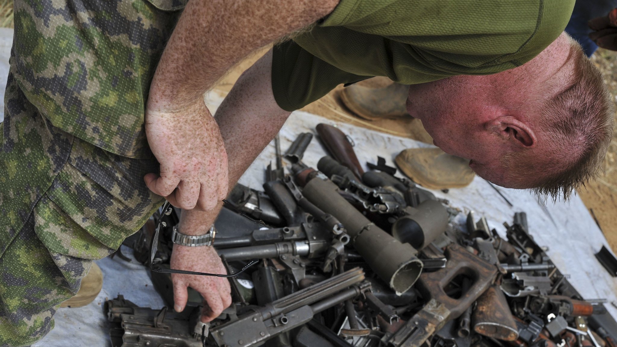 Dans cette image, on peut voir un homme en tenue militaire, penché en avant, qui examine un tas d'armes. Ce tas, étalé sur une surface comme une bâche ou un sol dur, est composé de diverses armes à feu, incluant des fusils et des pistolets, qui sont éparpillés de manière désordonnée. L'homme, avec des cheveux courts et roux, semble concentré dans sa tâche d'inspection. L'environnement autour de lui pourrait évoquer un contexte militaire ou de désarmement.