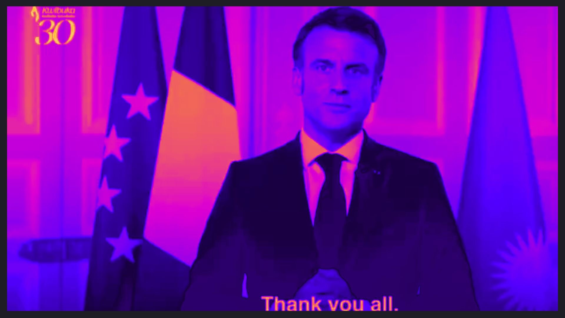 L'image montre un homme en costume sombre, debout devant un arrière-plan aux tons violets et roses. Il a une expression sérieuse et est placé entre deux drapeaux : un drapeau tricolore français à gauche et un autre qui pourrait représenter l'Argentine à droite. Ses mains sont jointes devant lui, comme s'il allait s'exprimer. En bas de l'image, on peut lire un texte en anglais qui dit "Thank you all", indiquant qu'il s'adresse à un auditoire. L'ensemble de la scène dégage une atmosphère formelle et solennelle.