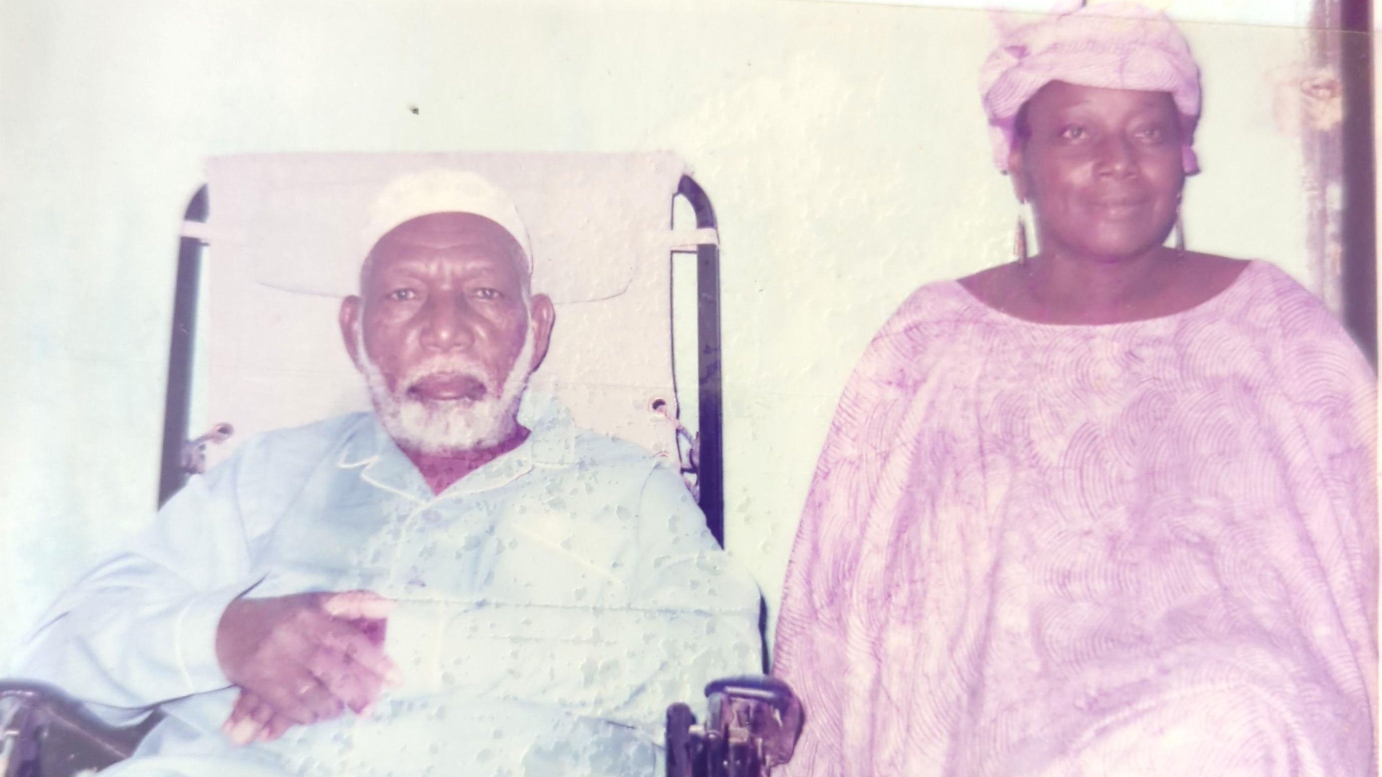 L'image montre un homme et une femme assis côte à côte. L'homme, plus âgé, est en fauteuil roulant et porte une tenue traditionnelle, comprenant un turban blanc. Il a une barbe grise et semble contemplatif. À côté de lui, la femme, qui a également des traits traditionnels, est vêtue d'une robe fluide et d'un foulard. Elle a une expression douce et chaleureuse. Le fond de l'image est sobre, créant un contraste avec leurs vêtements. L'atmosphère dégage une sensation de respect et de sérénité.