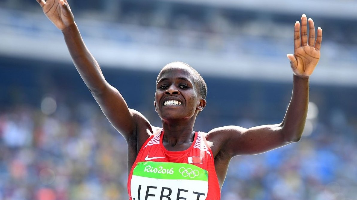 L'image montre une athlète célébrant sa victoire. Elle porte un débardeur rouge avec le nom "JEBET" inscrit dessus, et un logo des Jeux Olympiques sur sa tenue. Ses bras sont levés en signe de triomphe, et elle affiche un large sourire, exprimant la joie et l'excitation de son succès. En arrière-plan, on peut deviner une foule dans un stade, ajoutant à l'atmosphère festive de l'événement. L'éclairage est lumineux, mettant en valeur l'expression de bonheur sur son visage.