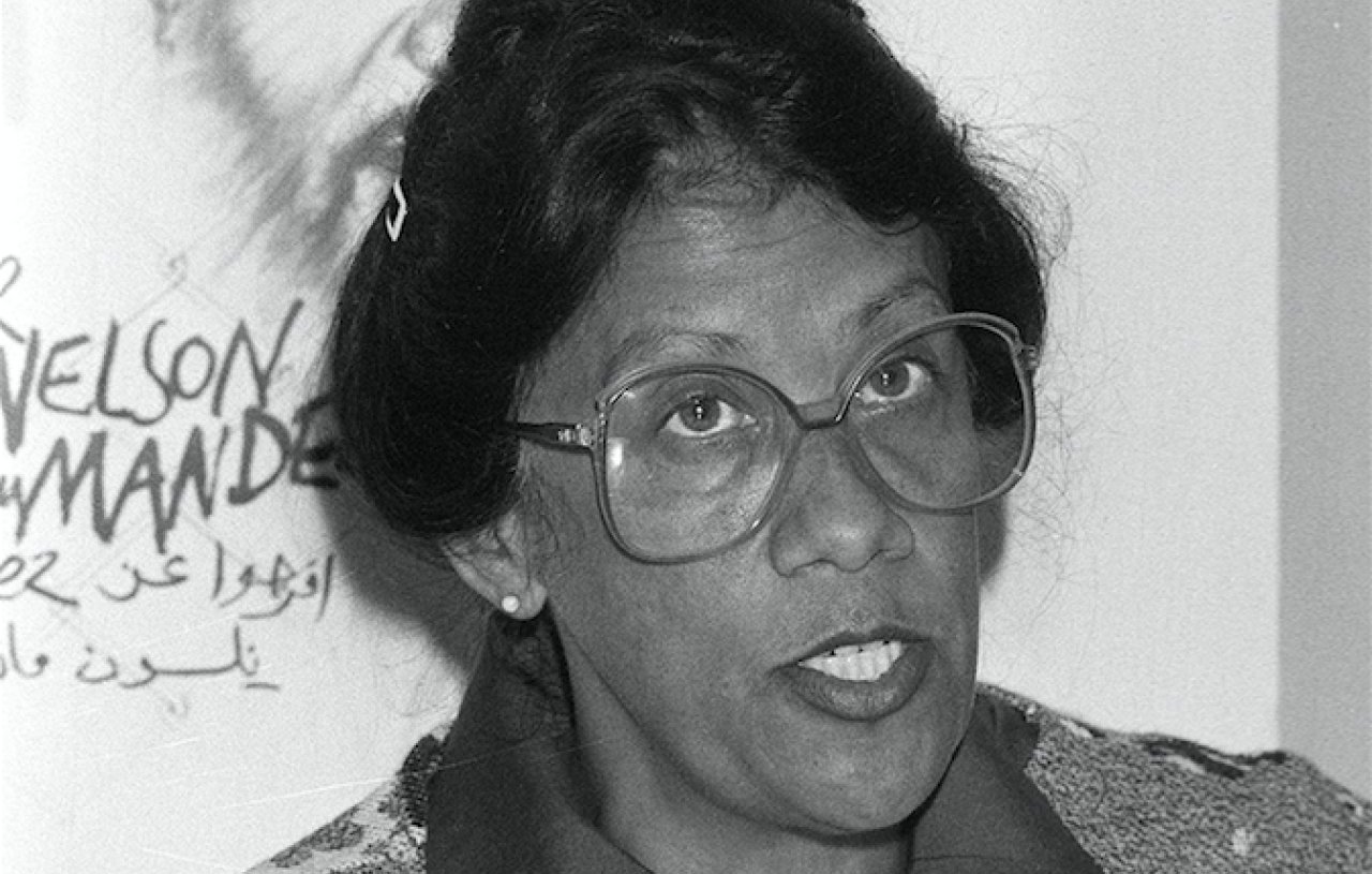 L'image montre une femme avec des lunettes épaisses, au visage expressif et aux cheveux noirs coiffés en un chignon. Elle porte un pull coloré avec un motif fleuri sur fond clair. En arrière-plan, on aperçoit une illustration d'un homme, probablement un portrait, qui ajoute un contexte à la scène. L'atmosphère semble sérieuse, et la femme semble engagée dans une discussion ou un discours. Ses traits et son regard transmettent une certaine détermination et une passion pour le sujet abordé.