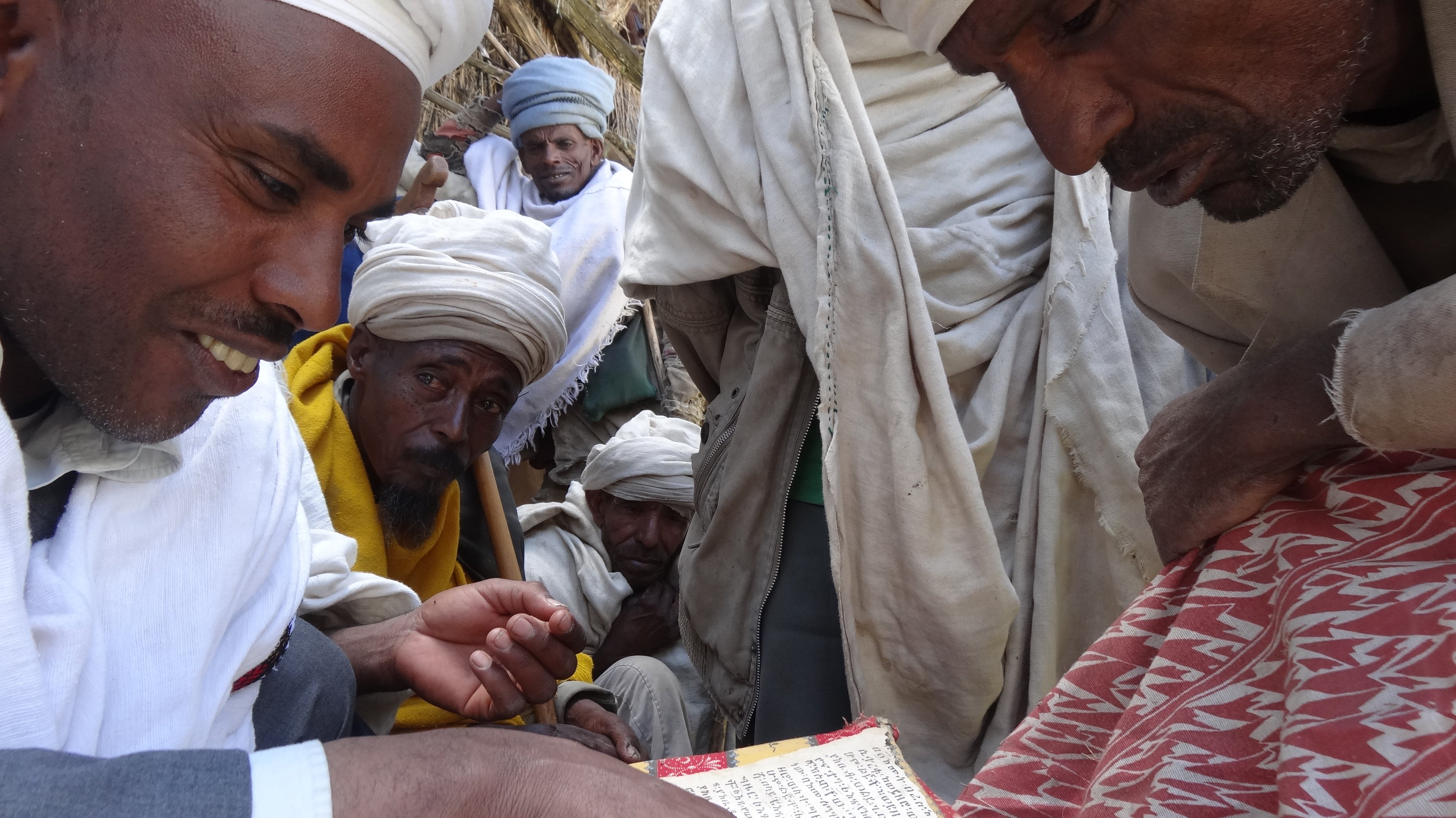 L'image montre un groupe de personnes, principalement des hommes, qui se rassemblent autour d'une table basse sur laquelle se trouve un ancien manuscrit. Un homme au premier plan, portant un habit traditionnel et un turban, montre du doigt une partie du texte. Les autres hommes, qui semblent intéressés, l'observent attentivement. Ils portent également des vêtements traditionnels, et leur expression trahit un mélange de curiosité et de respect. L'ambiance de la scène évoque un moment de partage et de transmission de connaissances, dans un cadre simple mais chargé de culture. En arrière-plan, des éléments naturels et des textures de tissus ajoutent une profondeur à l'atmosphère.