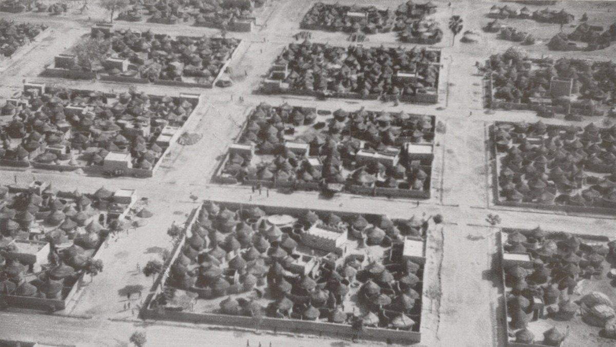 L'image montre une vue aérienne d'un village africain. On peut voir un agencement organisé des habitations, qui sont principalement des maisons traditionnelles en toit de chaume, caractéristiques de certaines régions. Les structures sont regroupées en plusieurs blocs délimités par des chemins. Des arbres sont dispersés dans l'espace, ajoutant une touche de verdure. Quelques personnes se déplacent le long des routes, suggérant une animation dans ce décor rural. Le photographique est en noir et blanc, ce qui donne une atmosphère historique à la scène.