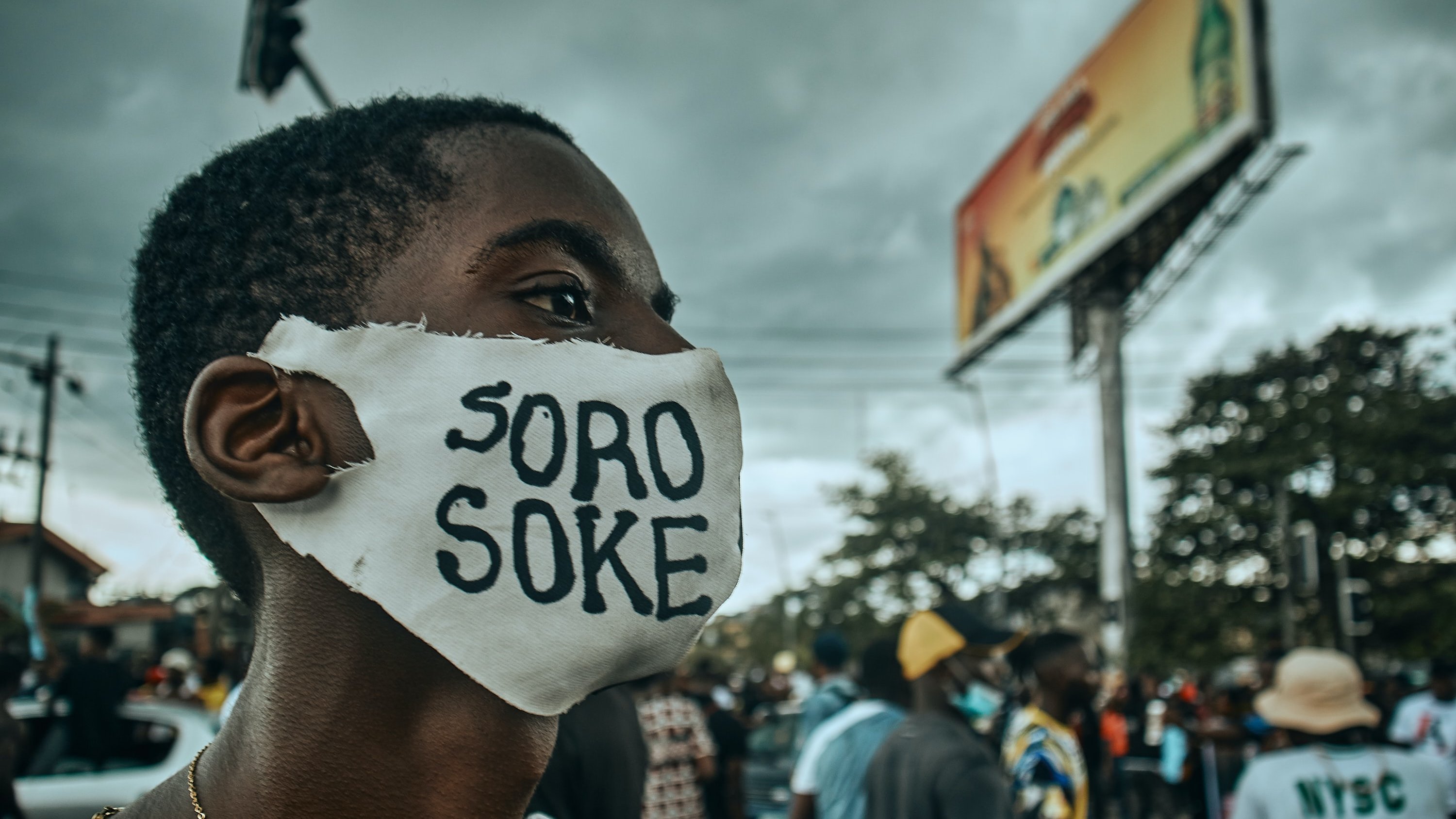 L'image montre une scène de manifestation dans une rue animée. Au premier plan, un jeune homme porte un masque blanc sur lequel est inscrit "SORO SOKE" en lettres noires, un slogan qui évoque la lutte pour les droits et la justice. Son regard est déterminé, et on peut sentir l'énergie de la foule qui l'entoure. À l'arrière-plan, des silhouettes de personnes se déplacent, et des affiches publicitaires peuvent être aperçues. Le ciel est nuageux, ce qui ajoute une atmosphère de tension à la scène. L'ensemble dégage une forte impression d'engagement et de solidarité.