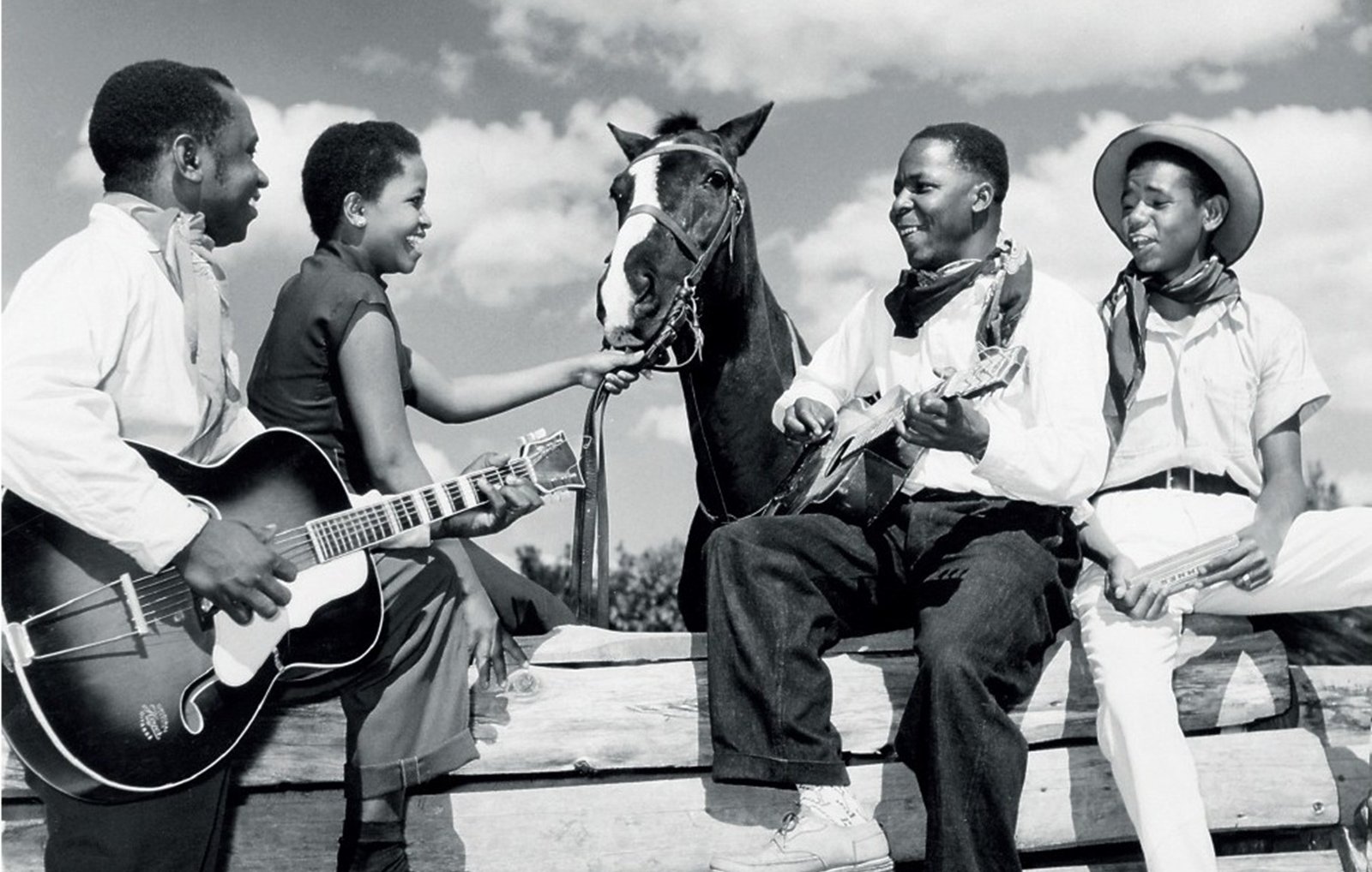 L'image représente un groupe de quatre personnes en noir et blanc, assises autour d'une barrière en bois, sous un ciel partiellement nuageux. Deux hommes et deux femmes sont présents. L'un des hommes joue de la guitare, tandis que l'autre est assis sur un tronc, tenant un instrument à cordes, probablement un banjo ou un ukulélé. Les deux femmes semblent interagir avec l'un des hommes et un cheval qui est également présent, la tête tournée vers eux. Elles affichent des expressions joyeuses, créant une atmosphère conviviale et détendue. Les vêtements des personnages varient : l'un porte un chapeau de cowboy et les autres portent des vêtements simples, typiques de l'époque. L'image évoque un moment de camaraderie et de musique, suggérant un lien fort entre les personnes et la nature environnante.